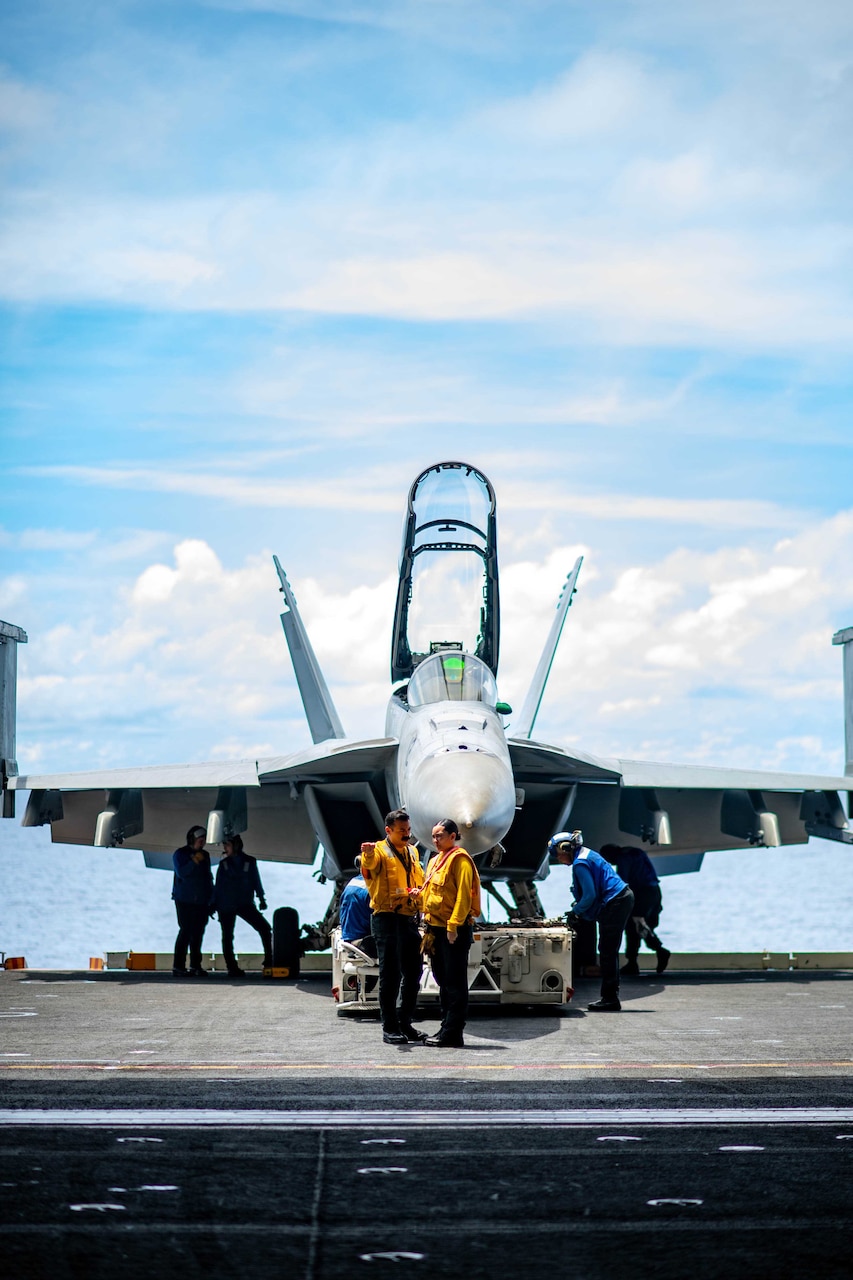 Sailors move supplies in front of an aircraft aboard a ship.