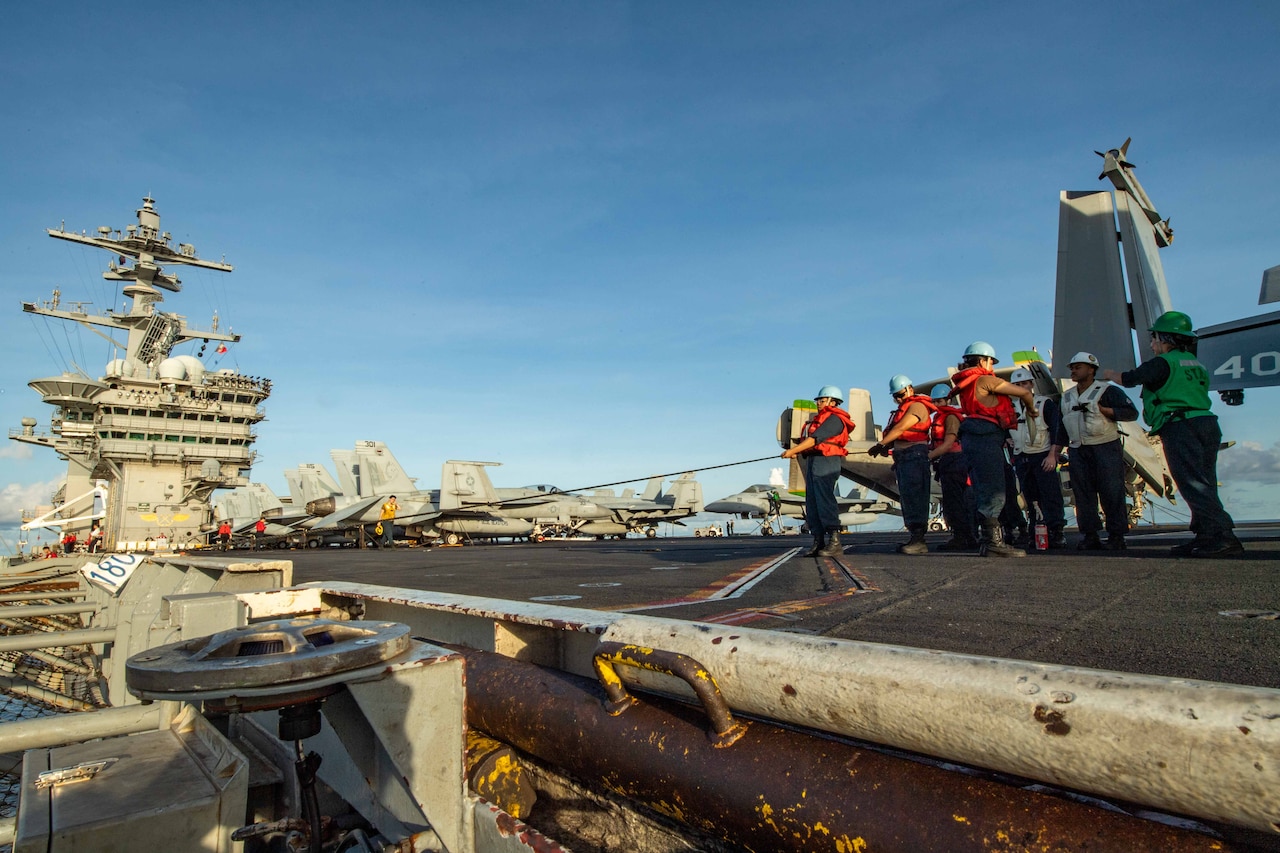 Sailors pull a line aboard a ship with aircraft in the background.