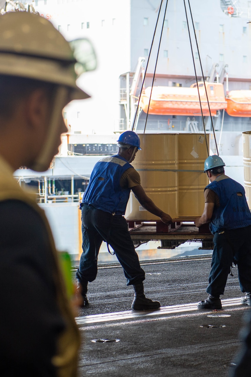 Sailors move cargo aboard a ship as fellow sailors watch.