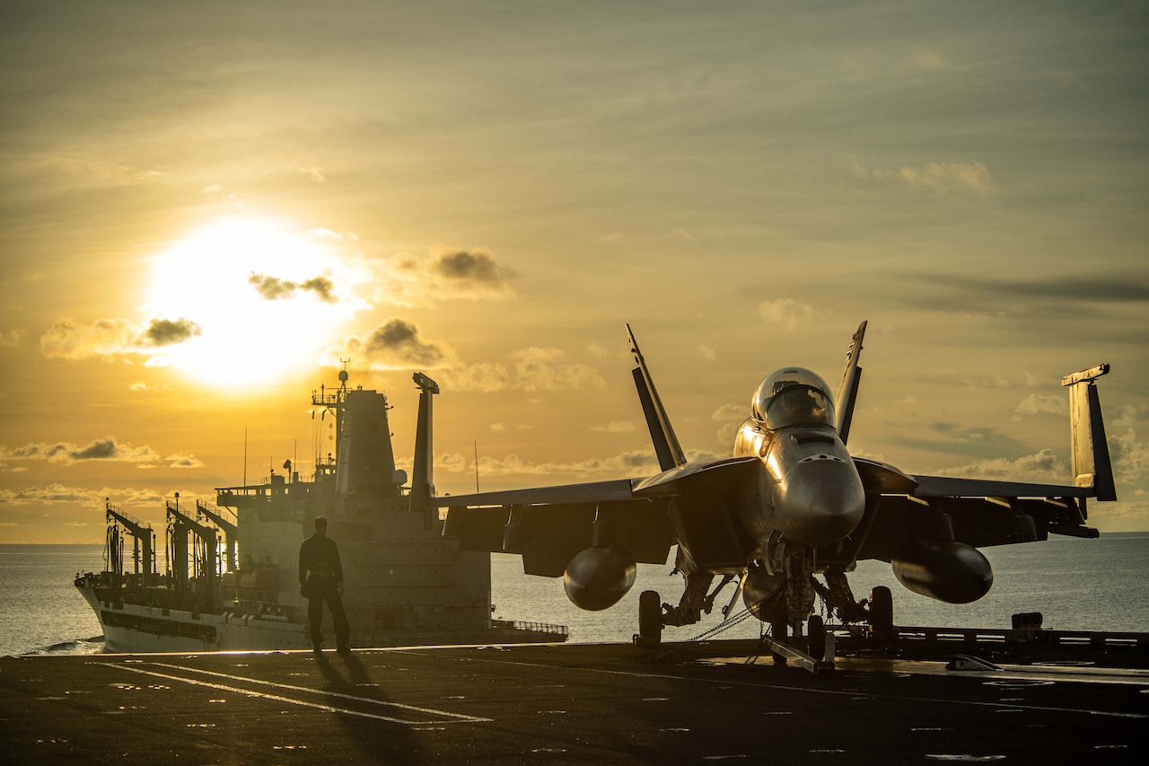 A sailor stands next to an aircraft aboard a ship at sea as another ship approaches under a sunlit sky.
