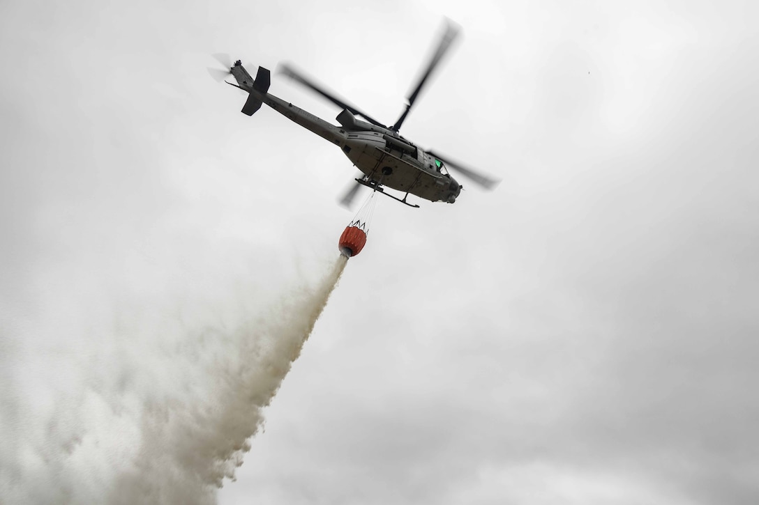 A helicopter seen from below flying in a cloudy sky uses a bucket to drop water.