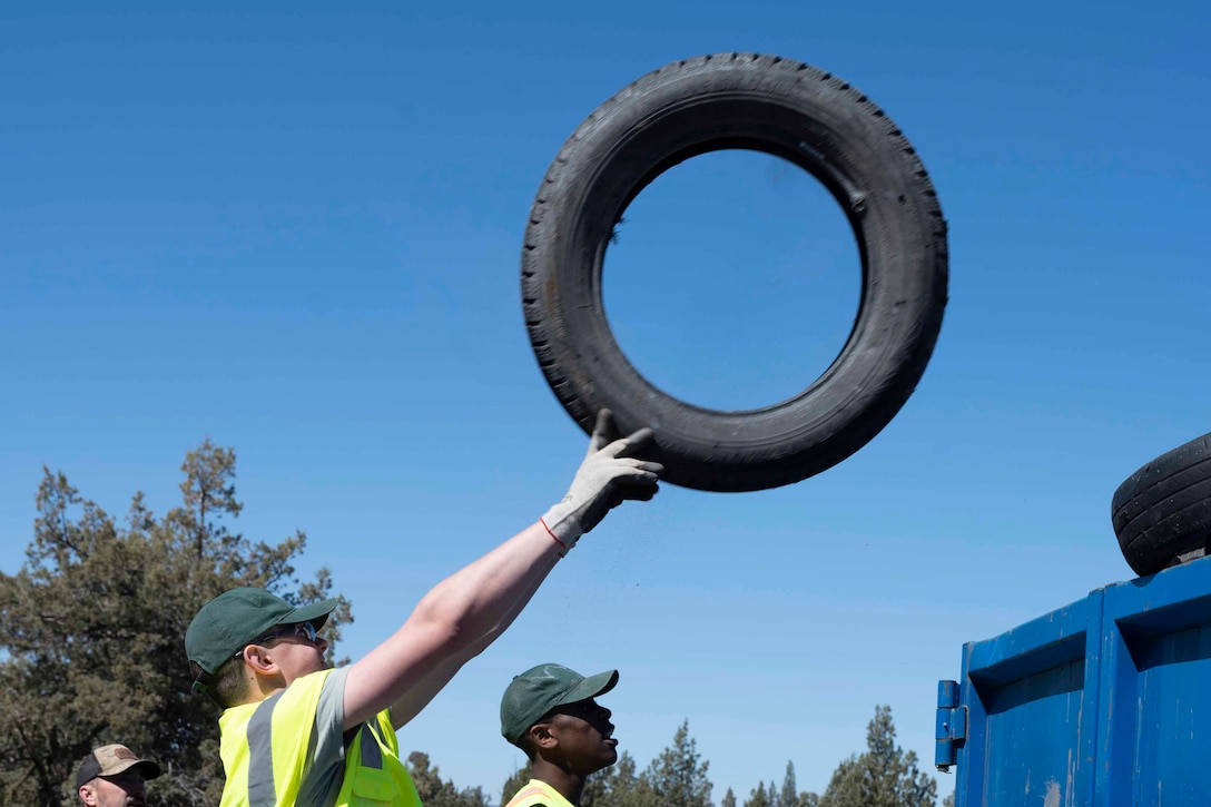 A volunteer tosses a tire into a dumpster under a bright blue sky while other volunteers watch.