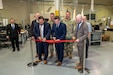 Eight men are pictured posing behind a red ceremonial ribbon tied across two short poles. Three of the men in the front are seen cutting the red ribbon.