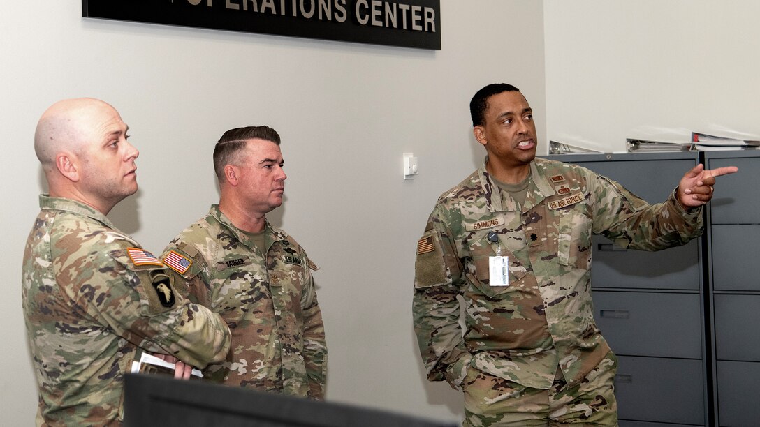 Three men stand against a wall in discussion