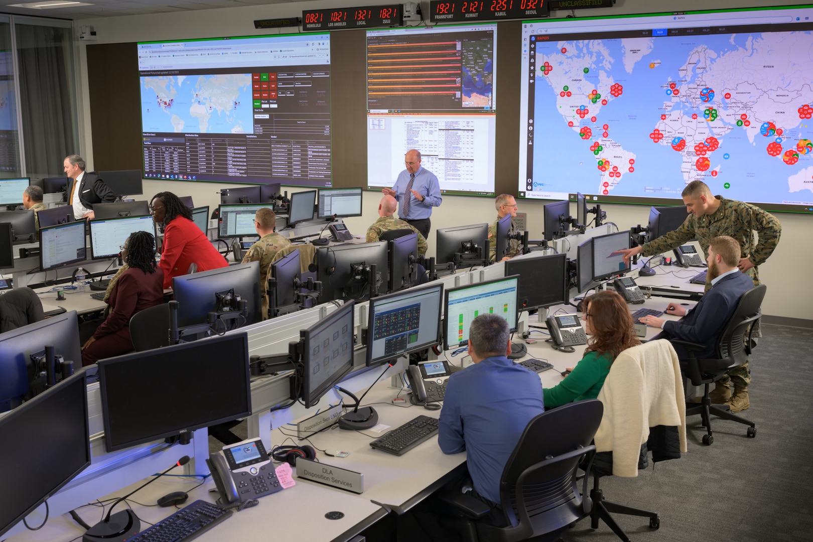 A group of people sit in front of computers or stand in a room with large monitors on the wall.