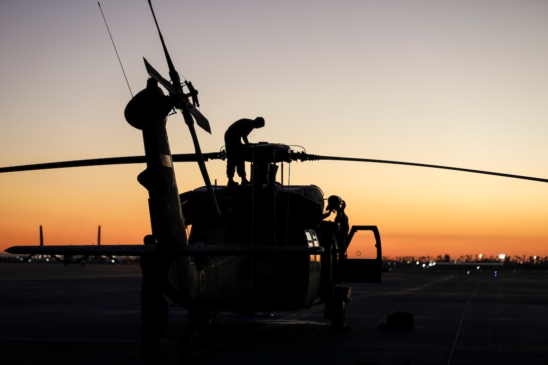 A service member is shown in silhouette standing atop a military helicopter at twilight against a fading orange sky.