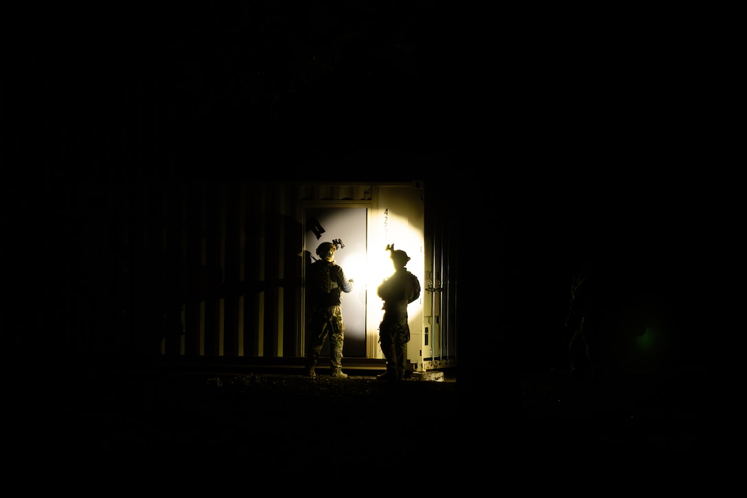 Two airman stand outside a door at night. They are illuminated by flashlights and headlamps.