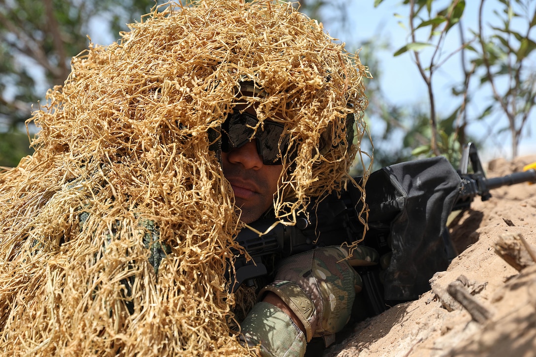 A service member covered with straw holds a weapon while hiding in a ditch during daylight.