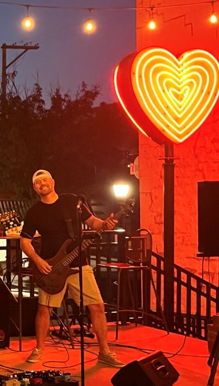 A man in a black shirt and khaki shorts holds a bass guitar with a yellow heart in the background.