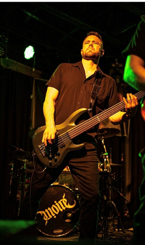 A man in a black shirt holds a black bass guitar.