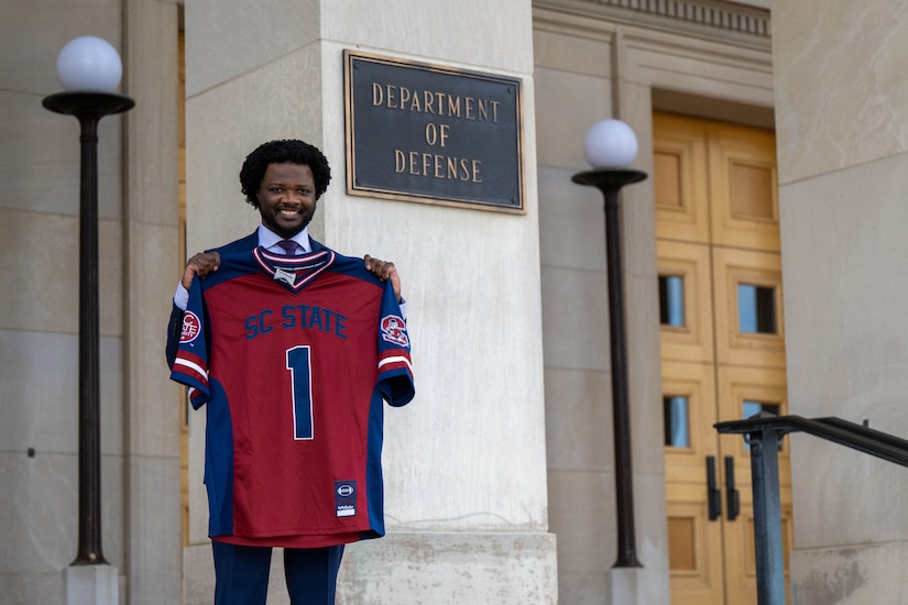 A person in a business suit holds up a jersey while standing in front of a building.