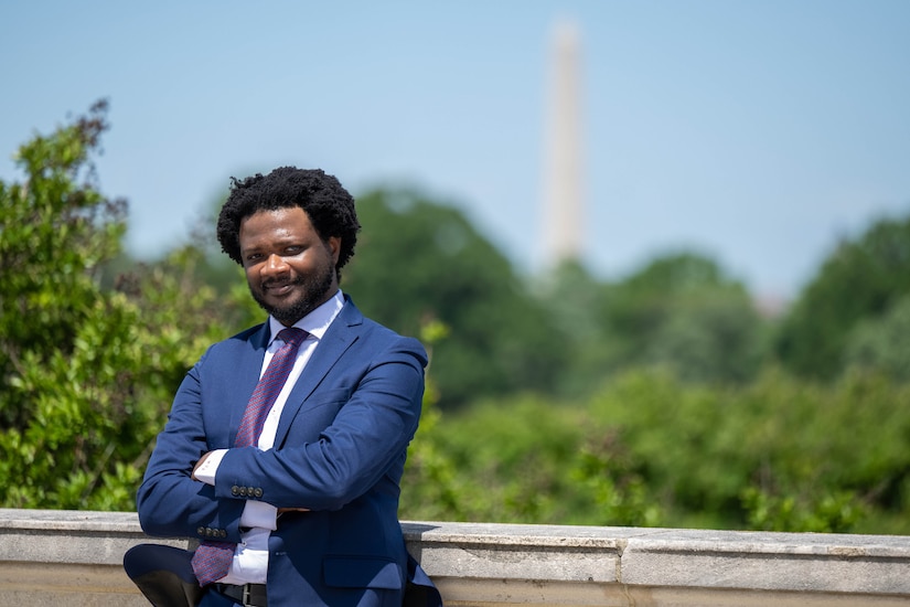 A person in a business suit stands near a building, and the Washington Monument is in the background.