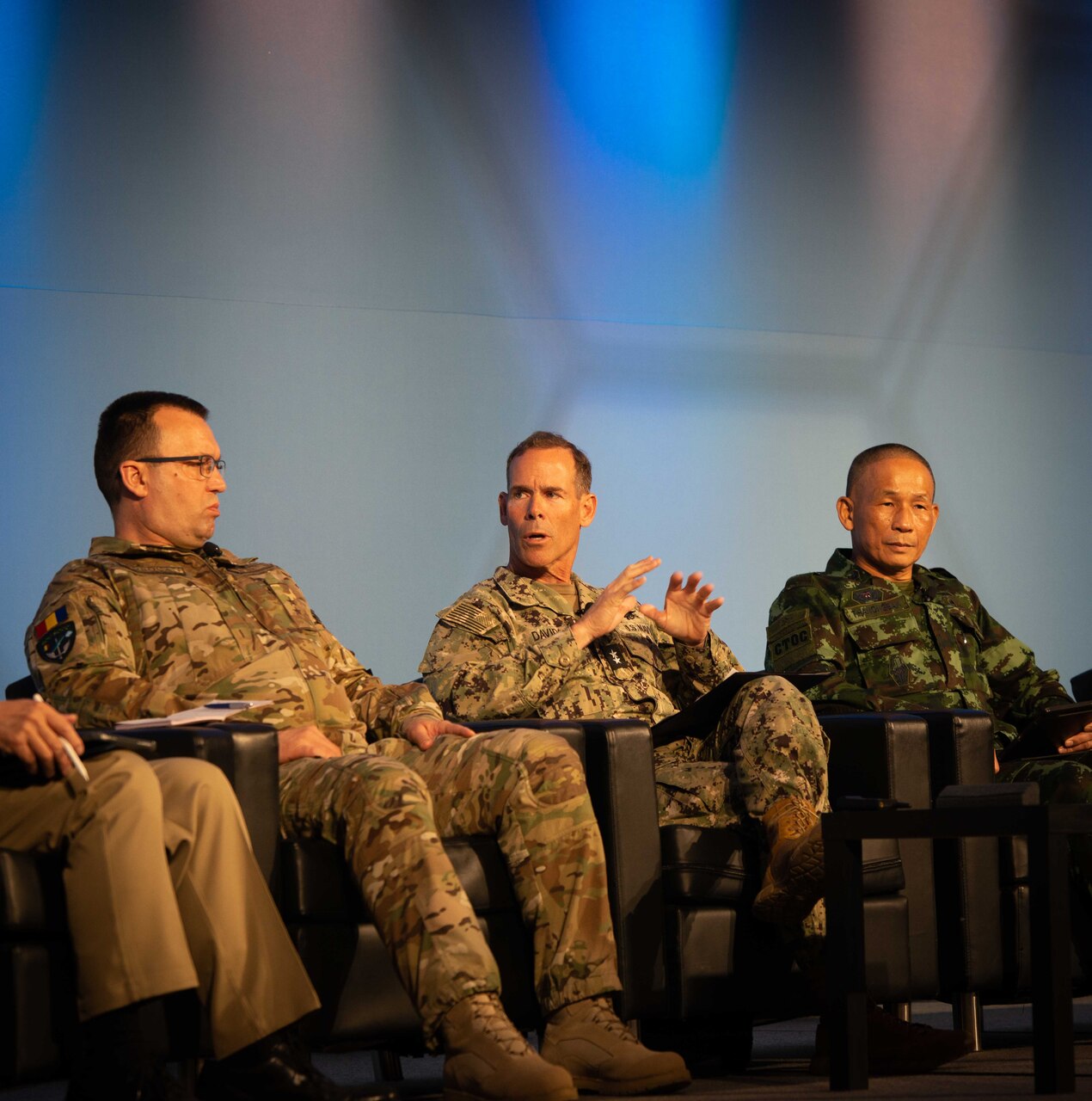 Three men in camouflage military uniforms sit in chairs. The man in the middle is looking to his right while gesturing with his hands.