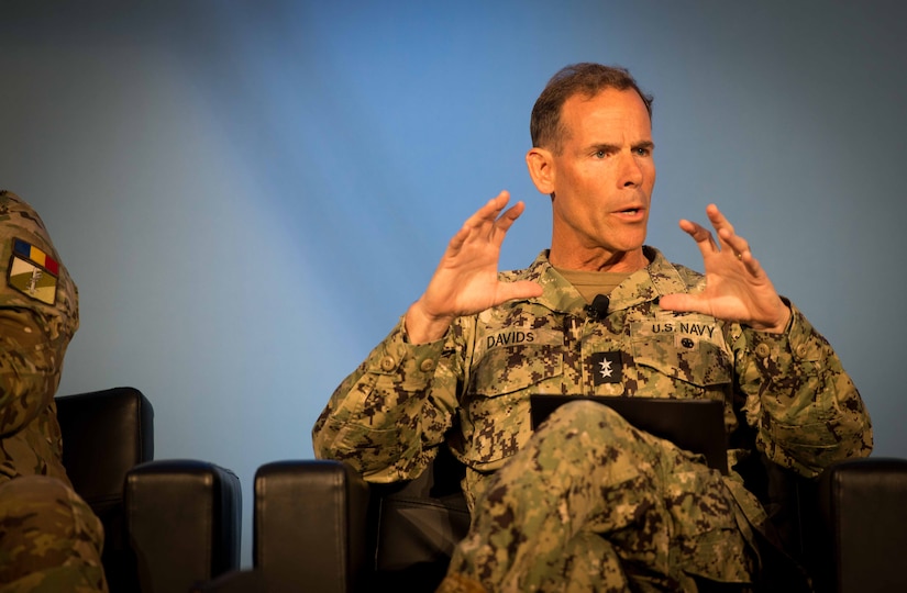 A man in a camouflage military uniform with two stars on the chest sits on a chair looking off screen while gesturing with his hands.