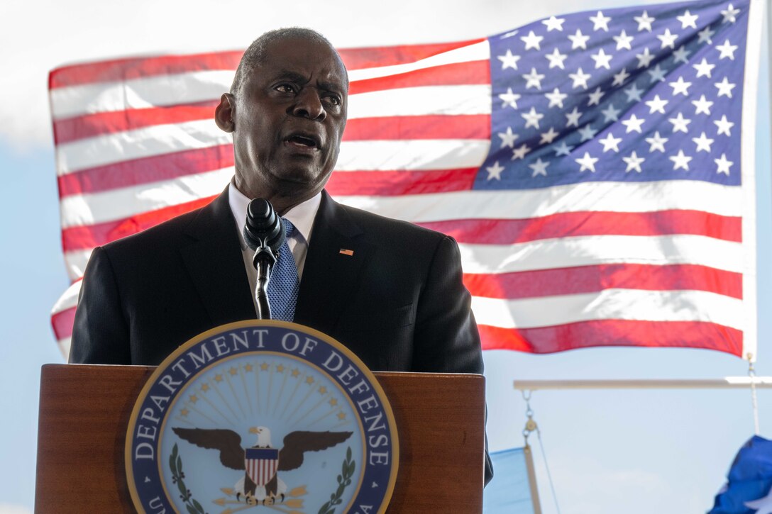 A civilian speaks from behind a podium with an American flag flying in the background.