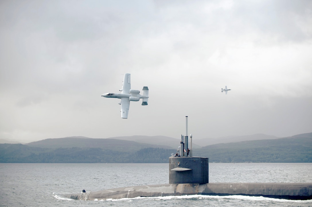 A submarine transits a body of water as two aircraft circle from above with mountains in the background.