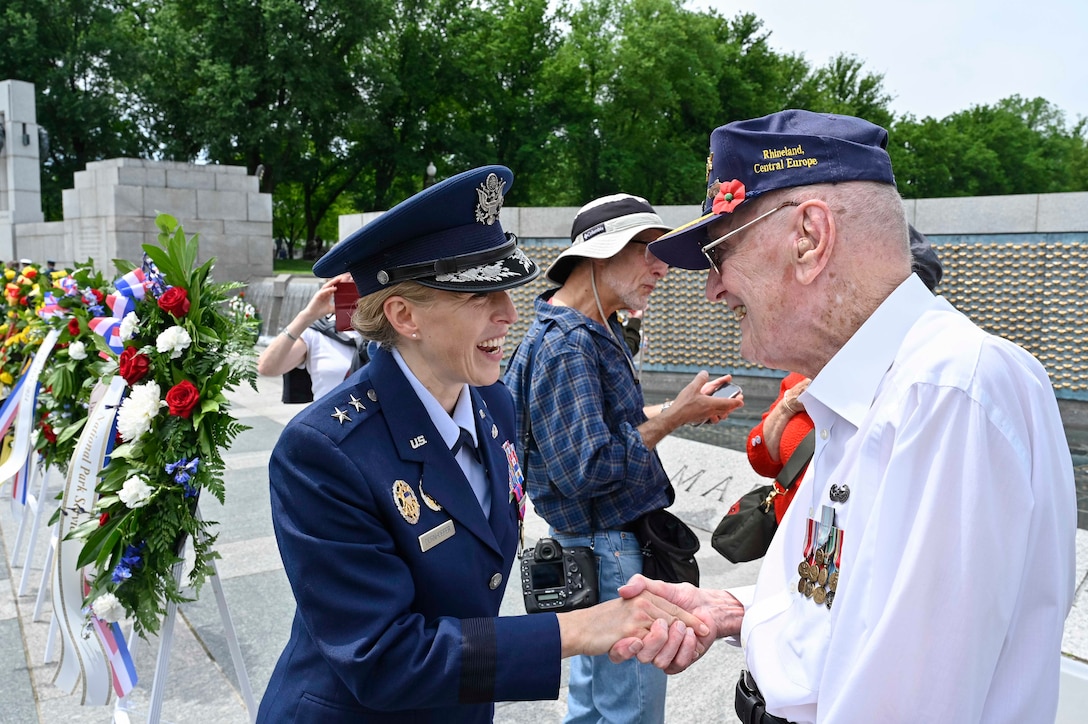 A military officer shakes hands with a veteran at the World War II Memorial with others and wreaths in the backdrop.