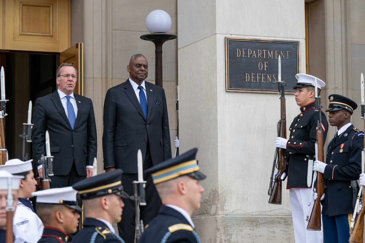 Two men stand together in front of a building. They are flanked by uniformed military personnel.  A sign notes that they are at the Department of Defense.