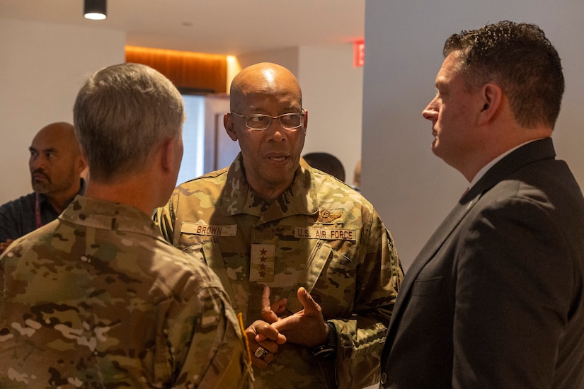 A person in a camouflage military uniform with four stars on it that reads “Brown” and “U.S. Air Force” on the chest talks with another person in a camouflage military uniform to their right and a second person in business attire to their left. The three are all inside a room.