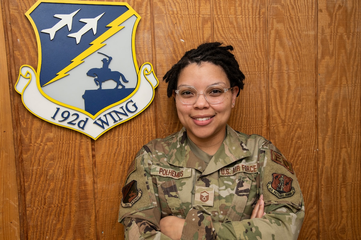 Master Sgt. Joclyn Polhemus stands with arms crossed next to 192nd Wing emblem.