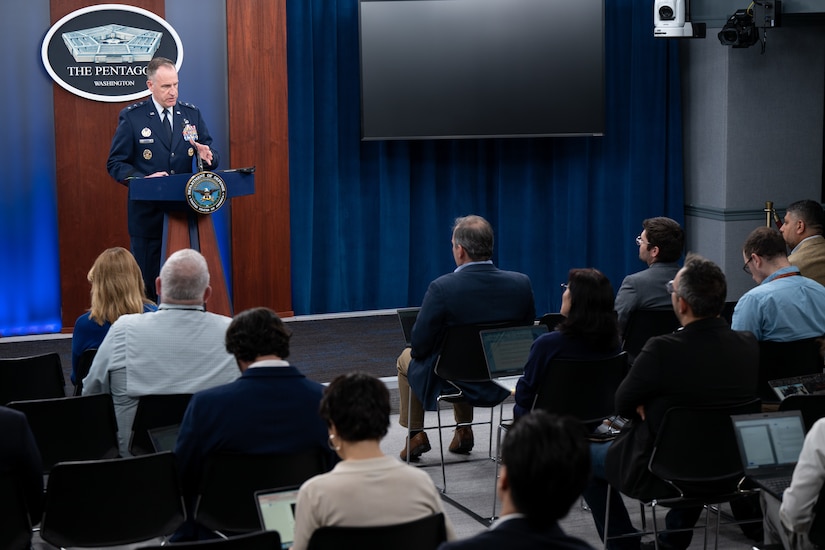 A man in a military uniform stands behind a lectern. Civilians are seated in front of him.