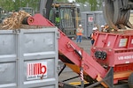 A shredder spits out wood fragments while personnel in safety gear observe.