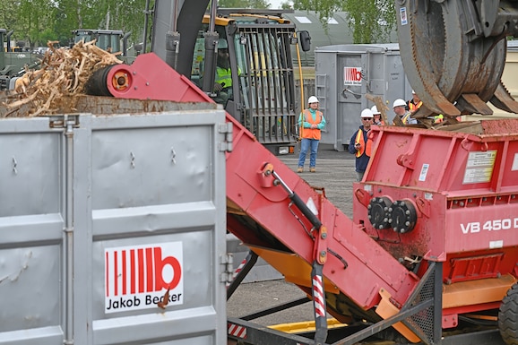 A shredder spits out wood fragments while personnel in safety gear observe.