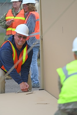 A man in safety gear strains while he tries to raise a foldable shelter wall.