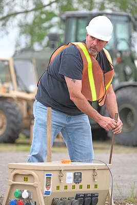 A man in safety gear prepares to drive a long metal stake into the ground.