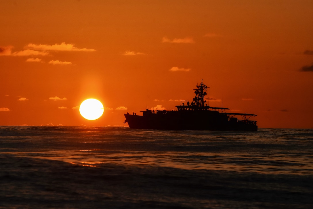 A ship is silhouetted against an orange sky and the sun as it moves across the water.