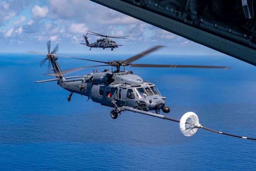 A helicopter receives fuel while in flight as another helicopter flies behind it.
