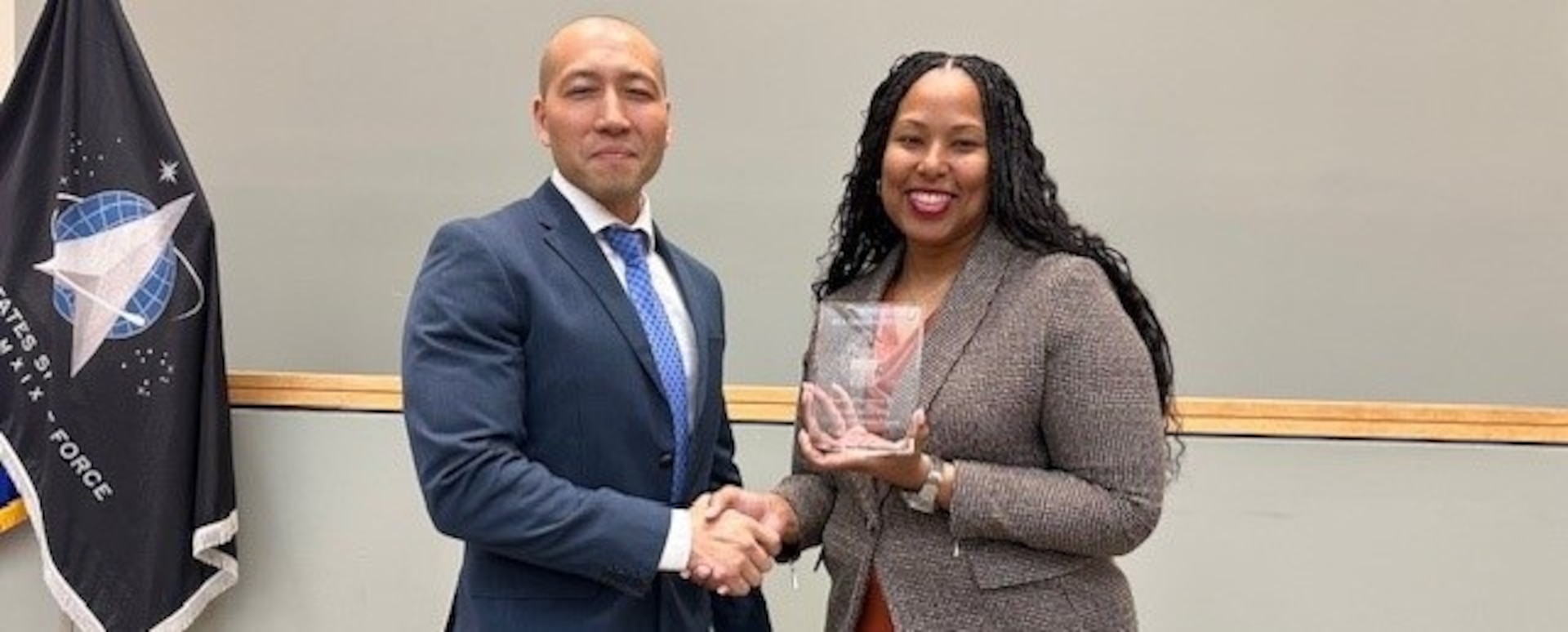 Kishayra Lambert presents Byung Kim a token of appreciation while standing in front of the Space Force flag.