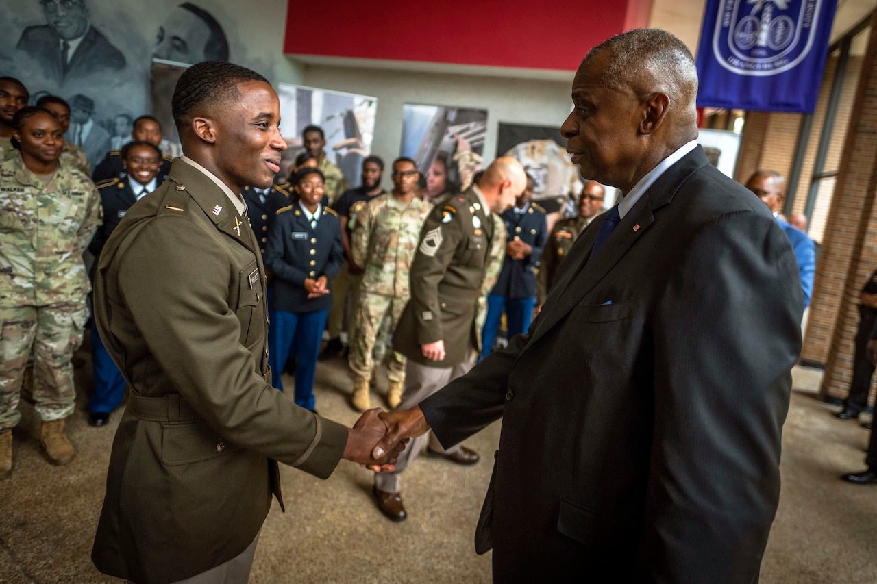 Secretary of Defense Lloyd J. Austin III shakes hands with a soldier in a lobby-type space with other troops in the background.