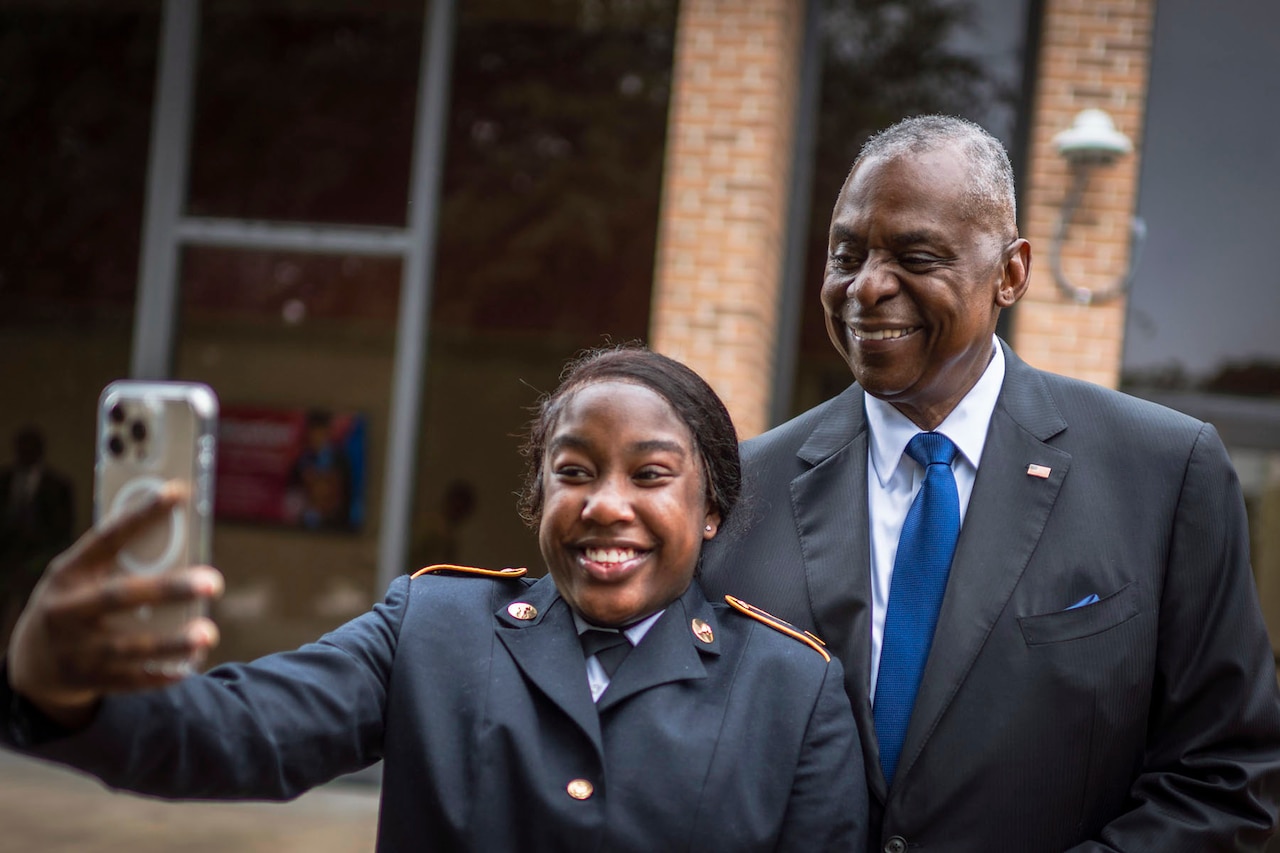 Defense Secretary Lloyd J. Austin III smiles while standing outside with a smiling ROTC cadet who holds up a phone for a selfie.