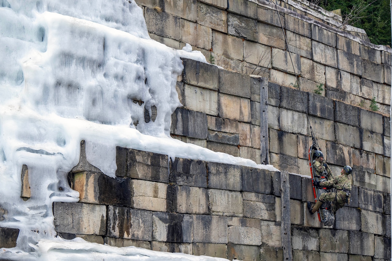 A man lowers a litter carrying another person down a vertical wall using ropes.