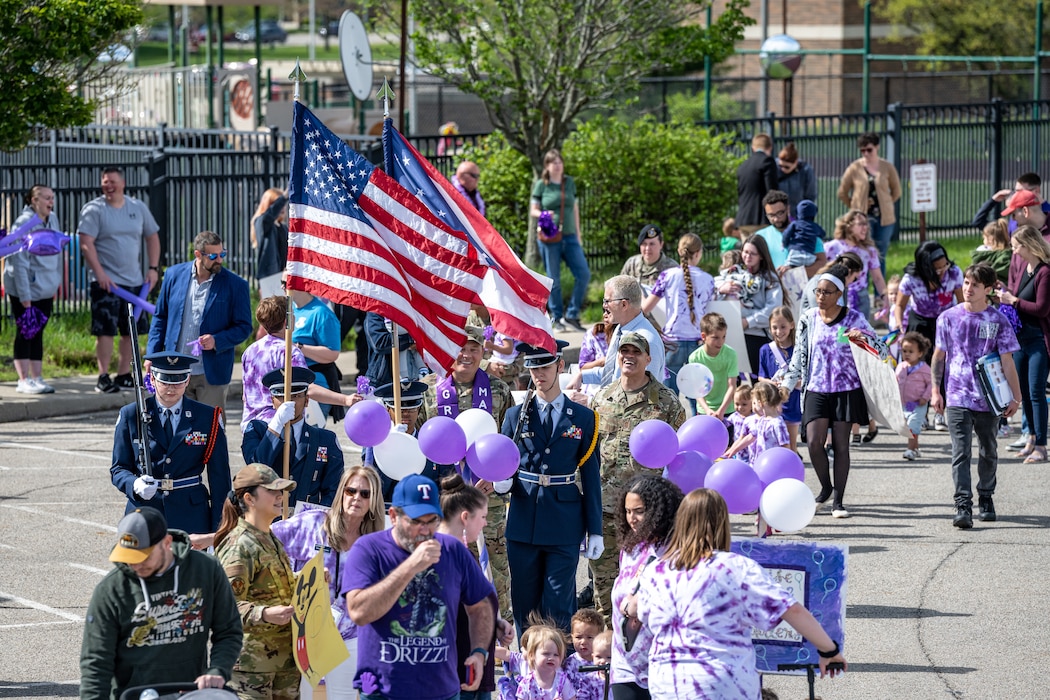 A wide image of a large procession of children in purple tie-dye, parents, servicemembers, and child care staff