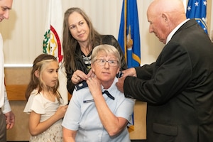 a woman is seated as another woman and man change the rank on her uniform shoulder board
