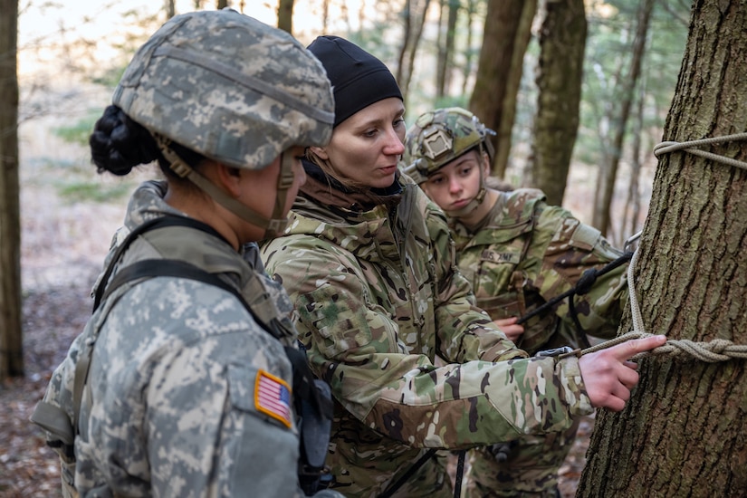 A woman points at a knot in a rope tied to a tree. Two other women look at it.