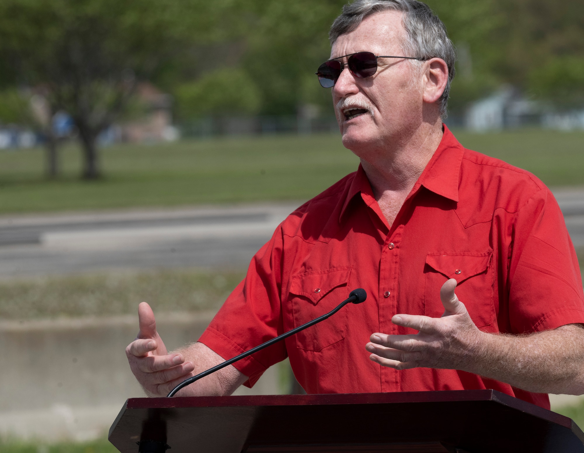 An older man in a red shirt gestures with his hands while speaking at a podium.