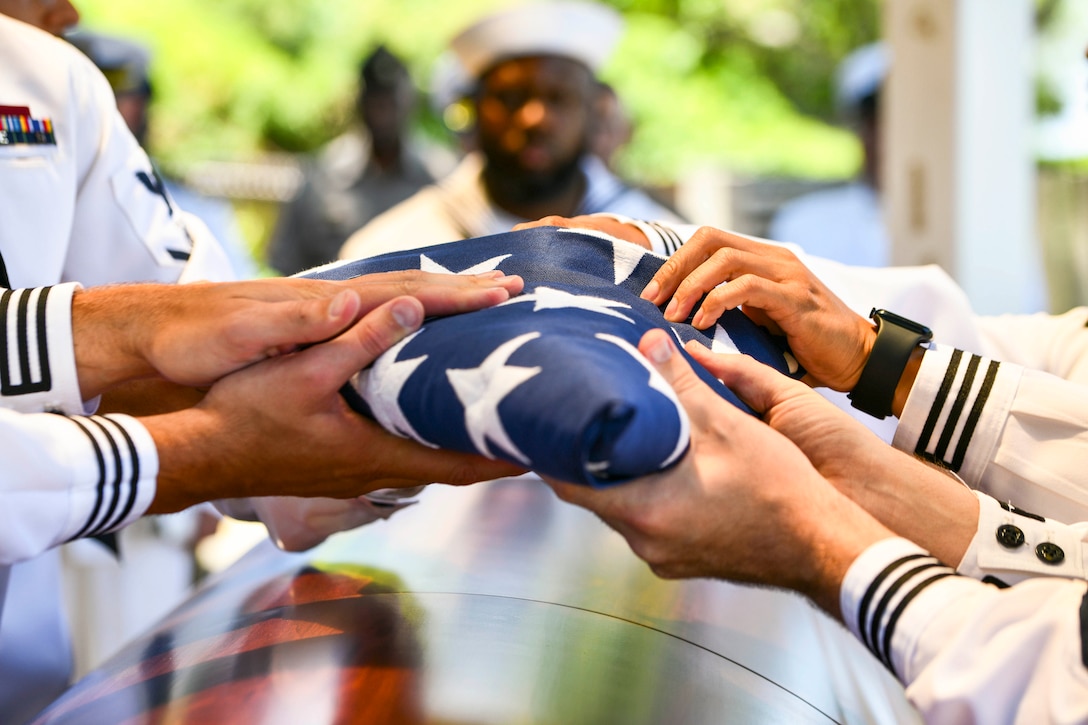 A close-up of sailors putting their hands on a folded American flag over a casket.