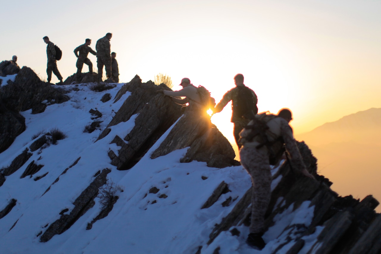 Several people walk the ridgeline of a snow-covered mountain as the sun lights them from behind.