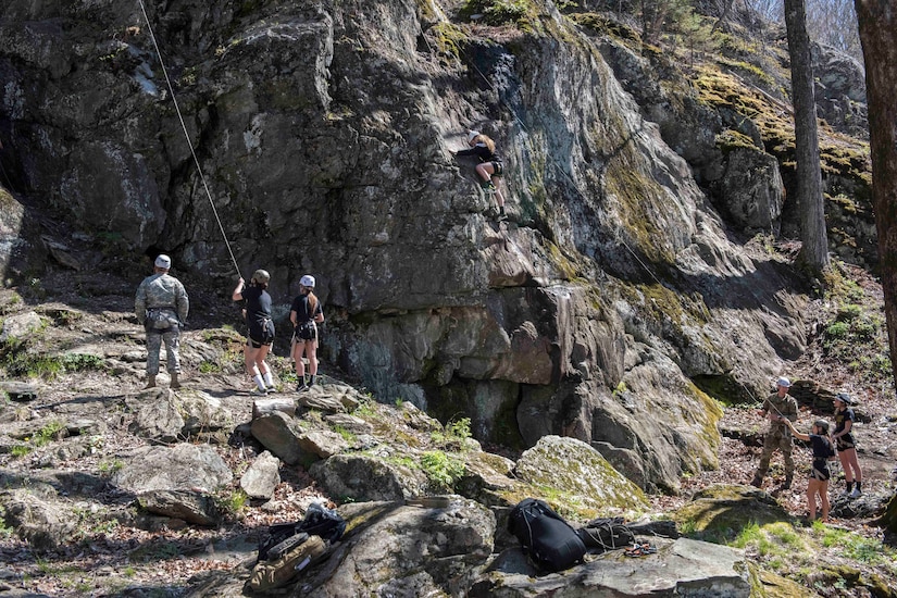 A man in uniform stands beside two other people, one of whom pulls a rope connected to the top of large mountain toward which they’re all looking.