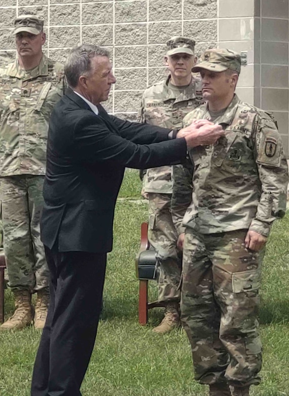 A soldier stands at attention while another man pins something to his lapel.