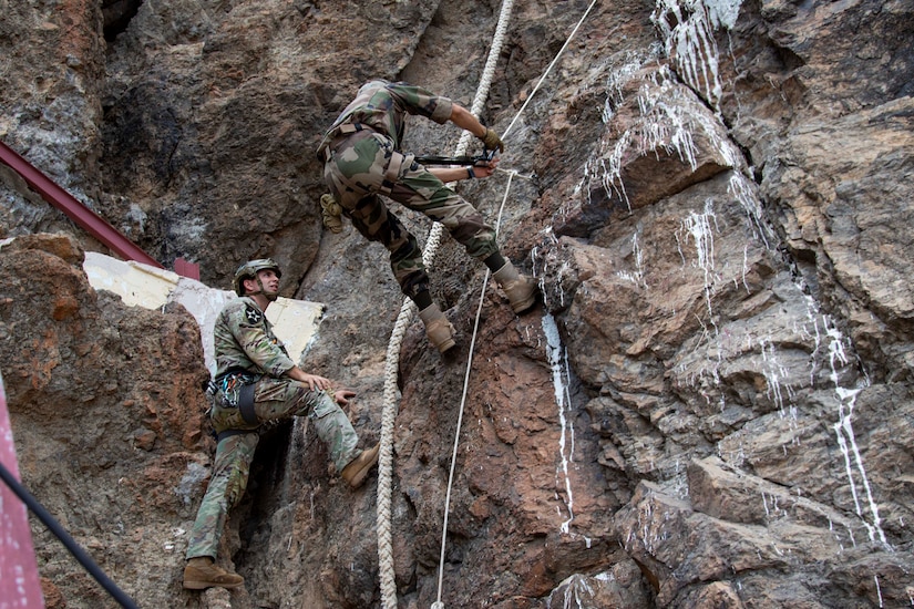 A man standing on the side of a rocky outcrop talks to another man scaling the vertical cliff.