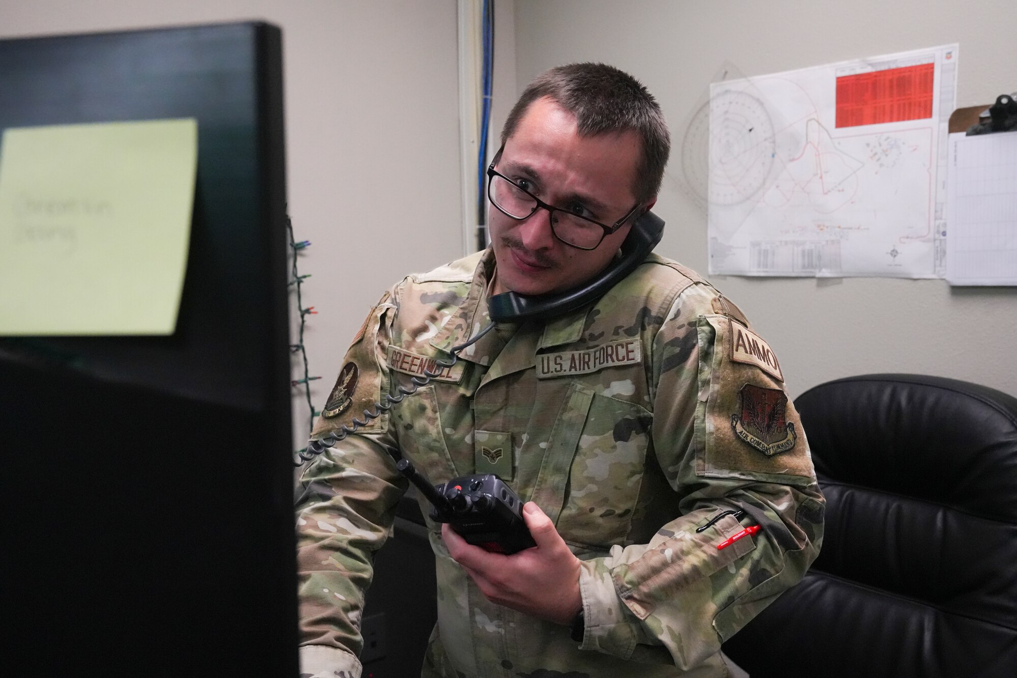 An airman on phone and holding a walkie talkie while looking at a computer monitor.