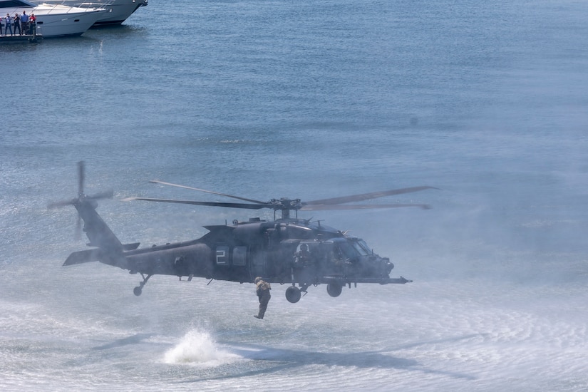 A man is jumping out of a hovering military helicopter approximately 15 feet to the water below.