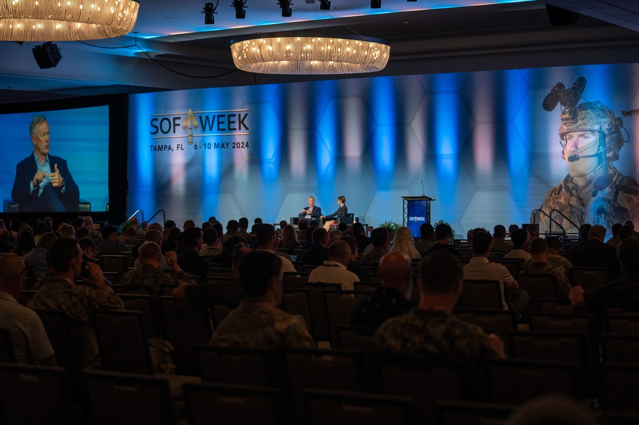 A man and woman are sitting down on a stage near a podium in a large conference room. A crowd is seated and observing them. To the left is a large projection of the man. The back wall has a picture of a service member in camouflage tactical gear. The wall reads “SOF Week, Tampa, FL 6-10 May 2024