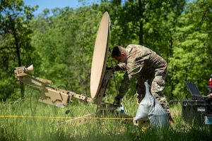 A U.S. Air National Guard Airman works to erect a portable satellite receiver. The receiver dish is in the midst of a wooded area.