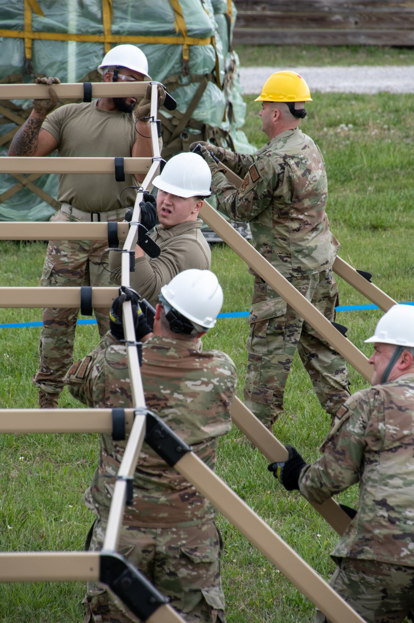 Several U.S. Air National Guard Airman work together to erect a large tent. The Airmen are wearing white safety hard hats.