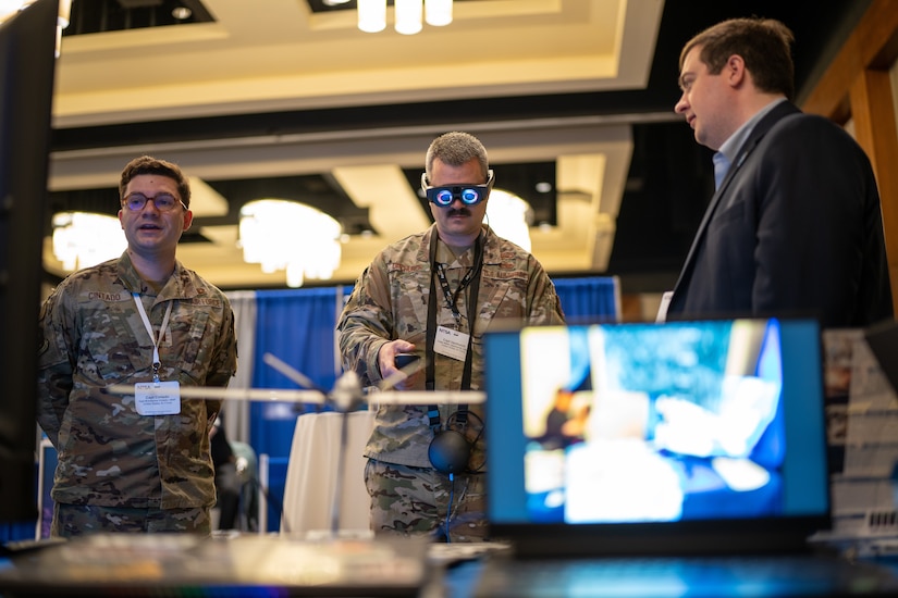 An airman wears goggles with tinted blue lenses while standing in a room near a monitor with another service member and a civilian.